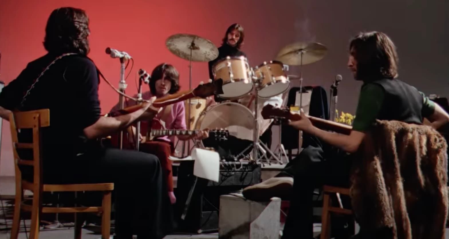 Three musicians rehearse in a studio, seated around a drum kit with guitars and warm red lighting.