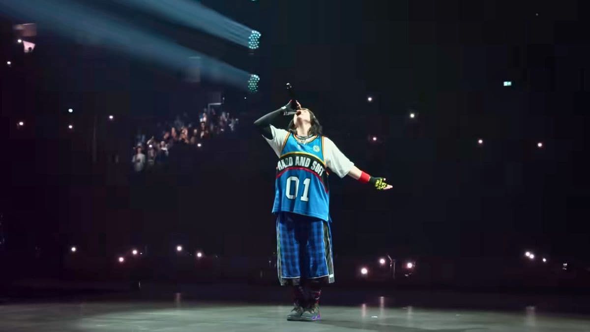 Performer in a blue jersey and hat standing on a dimly lit stage with spotlights and a crowd in the background