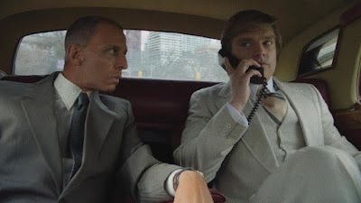 Two men in suits in a car. One on phone, the other looking on.