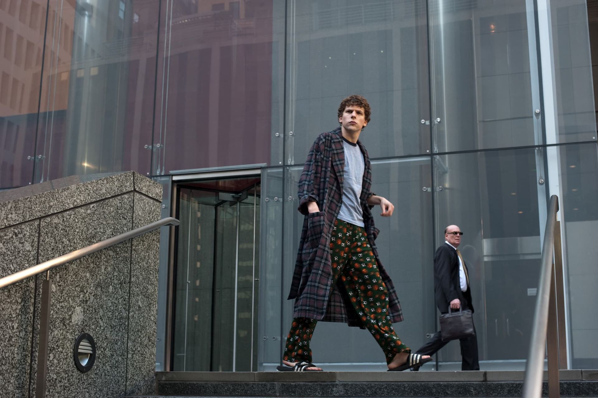 Man in a long dark coat walking outside a glass-fronted building, with another man behind him