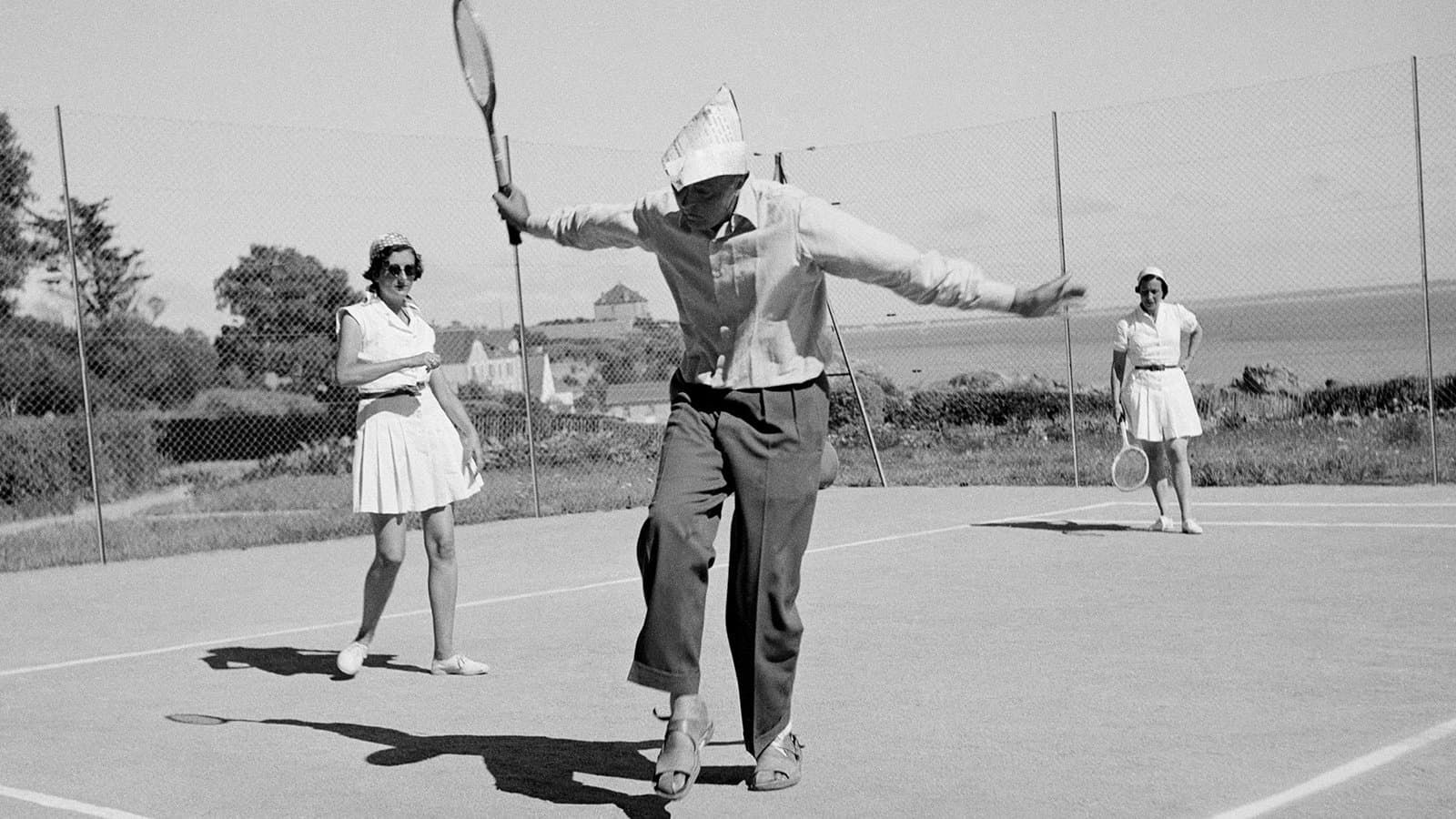Black-and-white photo of three people playing tennis on an outdoor court, one serving.