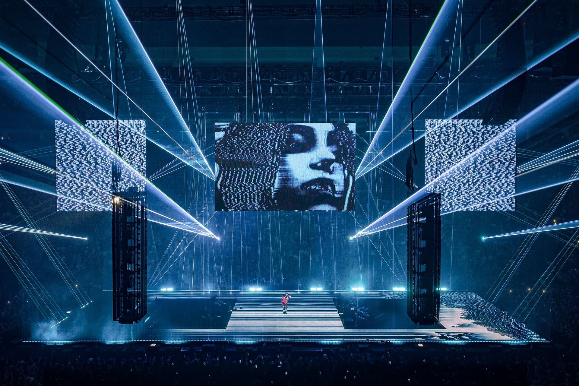 Blue-lit concert stage with a large screen, bright beams, and a lone performer at center stage