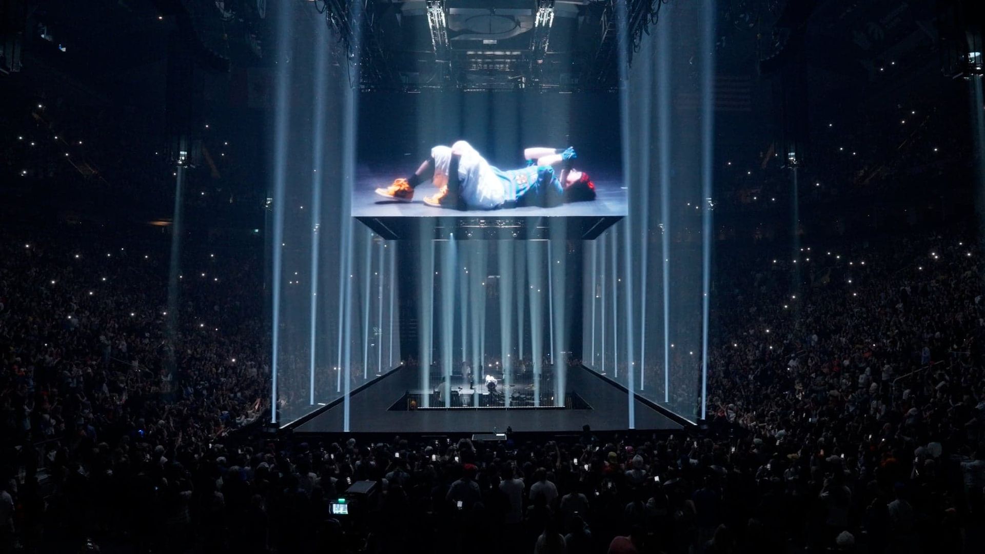 A performer lies on a glowing suspended platform above a dark concert crowd with vertical light beams.