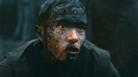 Close-up of a muddy, bloodied person staring tensely in a dark, wooded setting