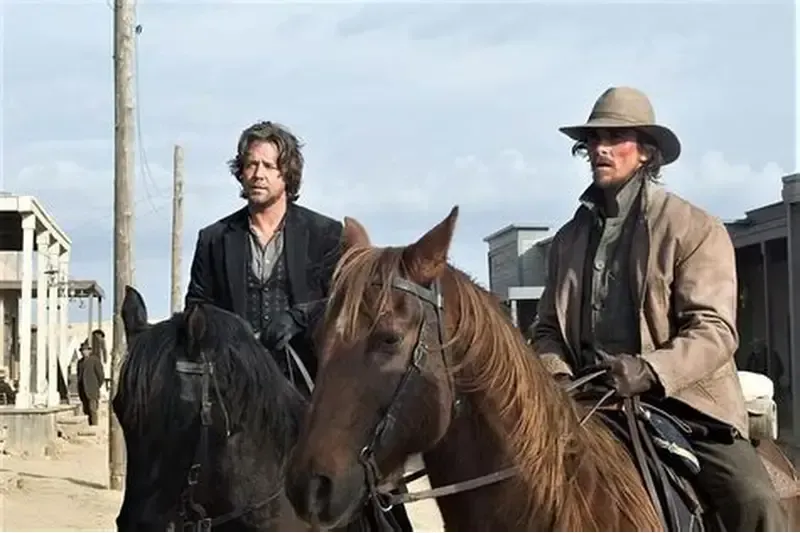 Two men on horseback in a dusty Western town. One wears a hat and brown coat, the other a dark suit.