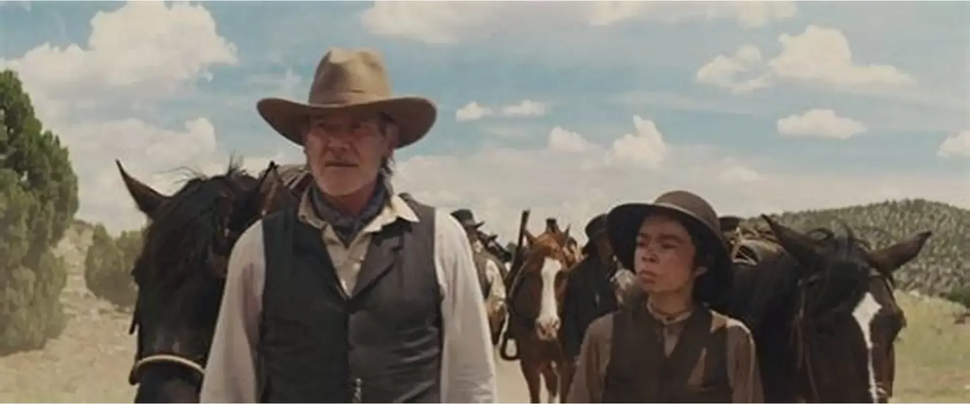 Two cowboys walking with a herd of horses on a dusty road under a cloudy sky.