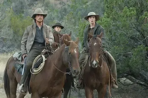 Three people on horseback in a dusty, outdoor setting. Man, boy, and teen ride brown horses.