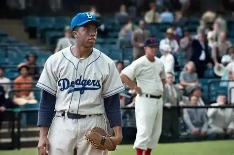 Chadwick Boseman as Jackie Robinson in a Dodgers uniform on a baseball field, looking determined.