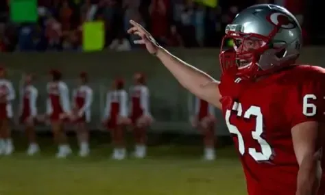 Football player in red uniform celebrates on the field, with cheerleaders in the background.
