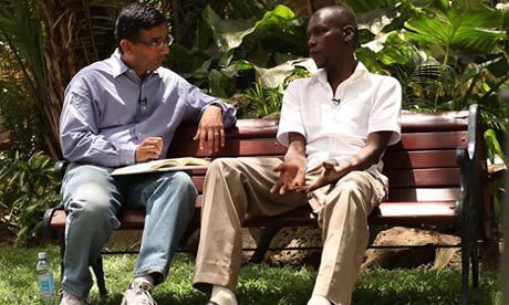 Two men on a park bench. One in blue shirt, one in white. Green foliage in background, sunny day.