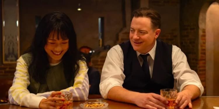 Two people laughing at a bar, seated with drinks in warm indoor lighting.