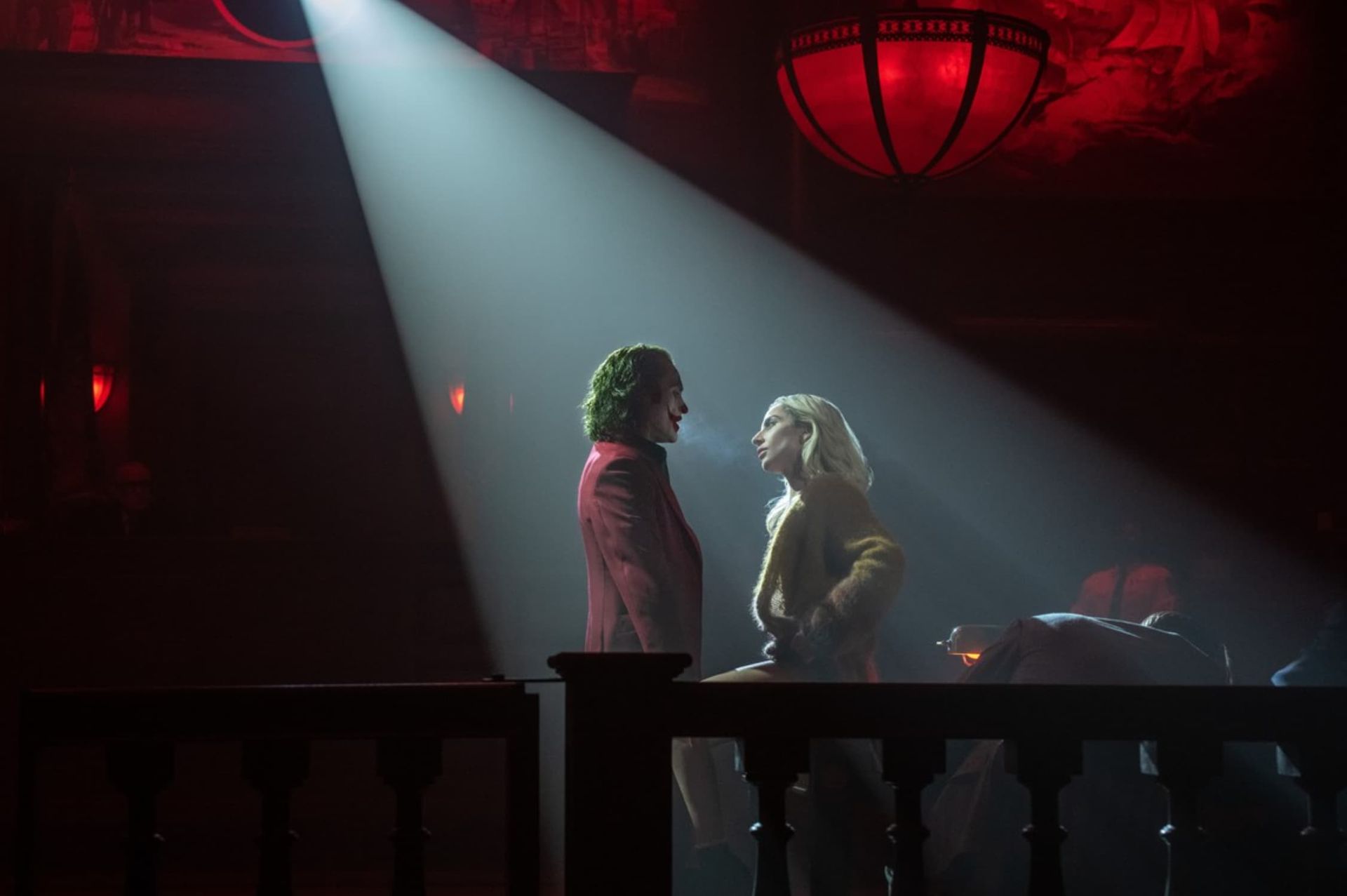 Joker and Harley Quinn stand face-to-face in a spotlight on a balcony, illuminated against a dark, red-lit background.