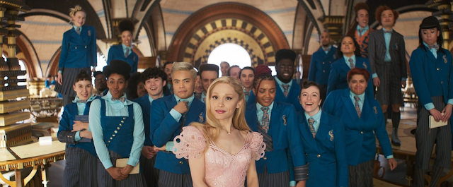 Blonde woman in a pink dress stands before a group in matching blue uniforms inside a grand hall