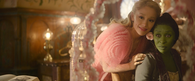 Two characters standing together indoors, one in a pink sweater and one with green face makeup.