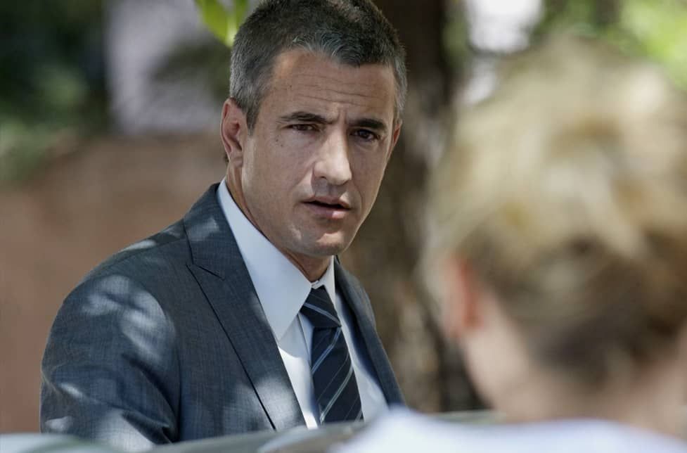 Man in a suit looking intently at a person off-screen. Outdoors, sunlight.