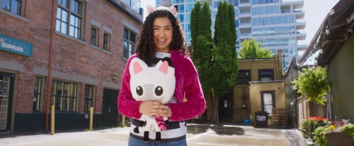 Woman in pink jacket and cat ears holds a plush toy, standing in an alley.