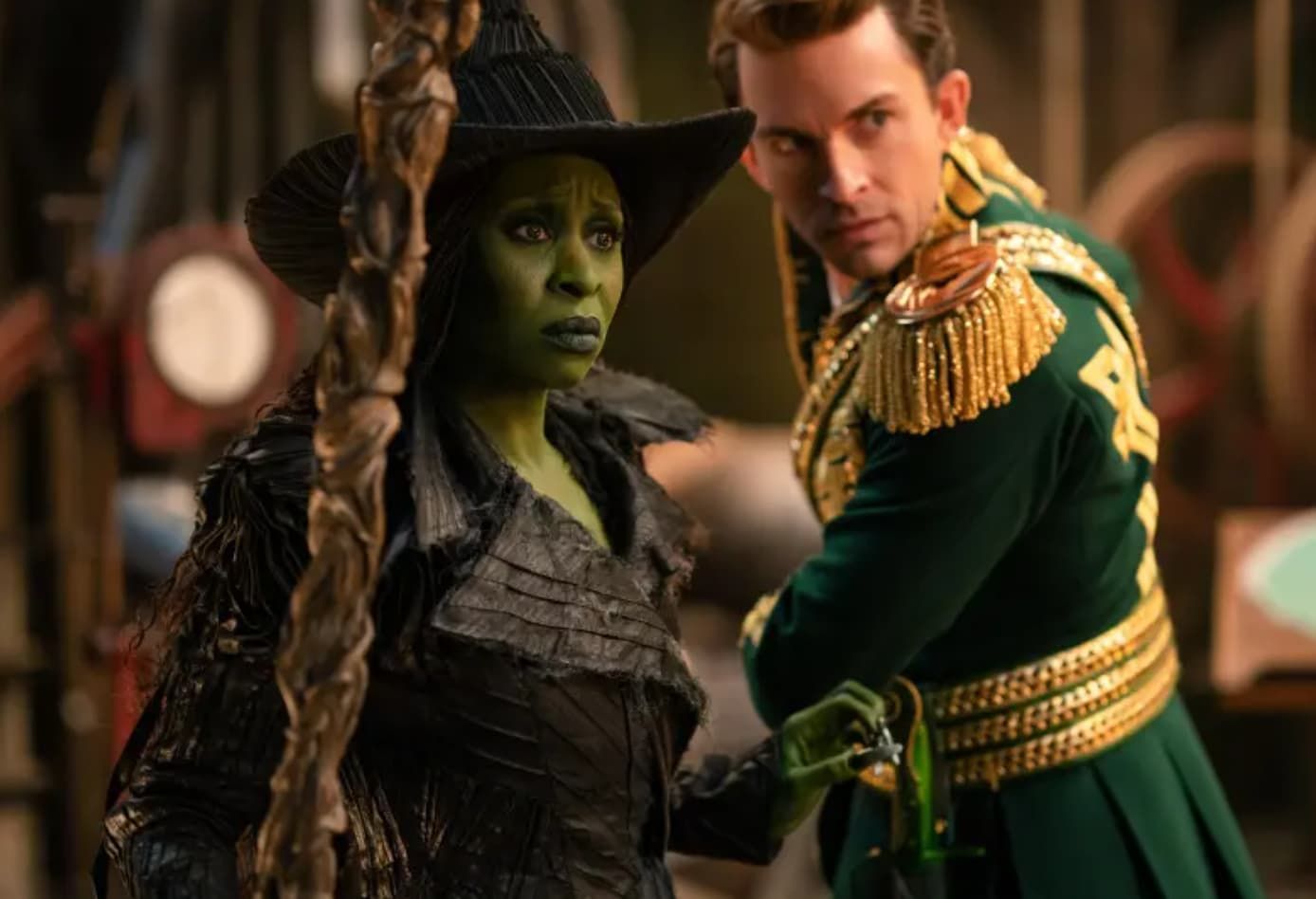 Green-skinned witch in a black hat and robe, looking concerned, next to a man in a green military-style jacket.