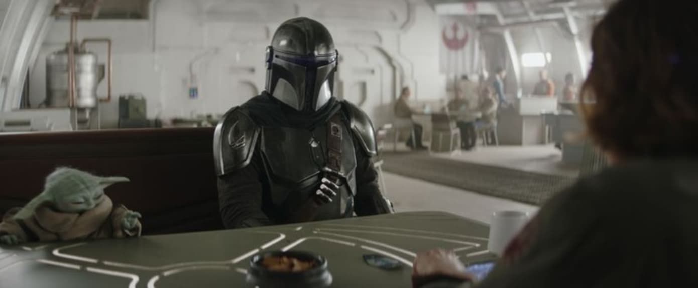 Mandalorian seated at a table in a bright room, facing a person across from him with a small green child nearby