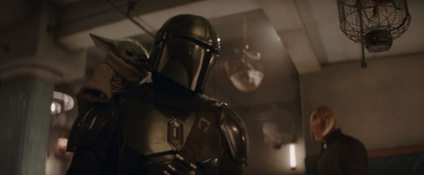 Armored Mandalorian stands in a dim room with a man in the background, warm light and hanging lanterns.