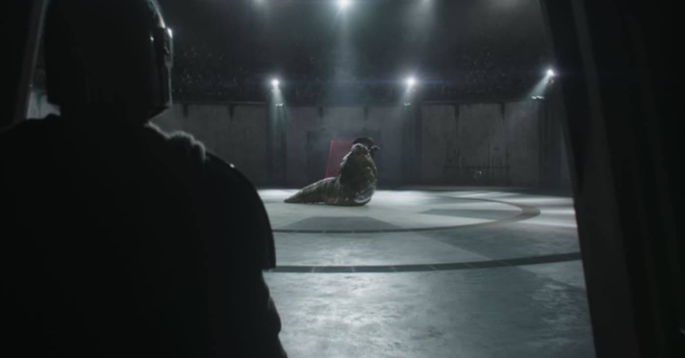 A dim stage with a spotlight on a seated figure, seen from behind a silhouetted person in the foreground.