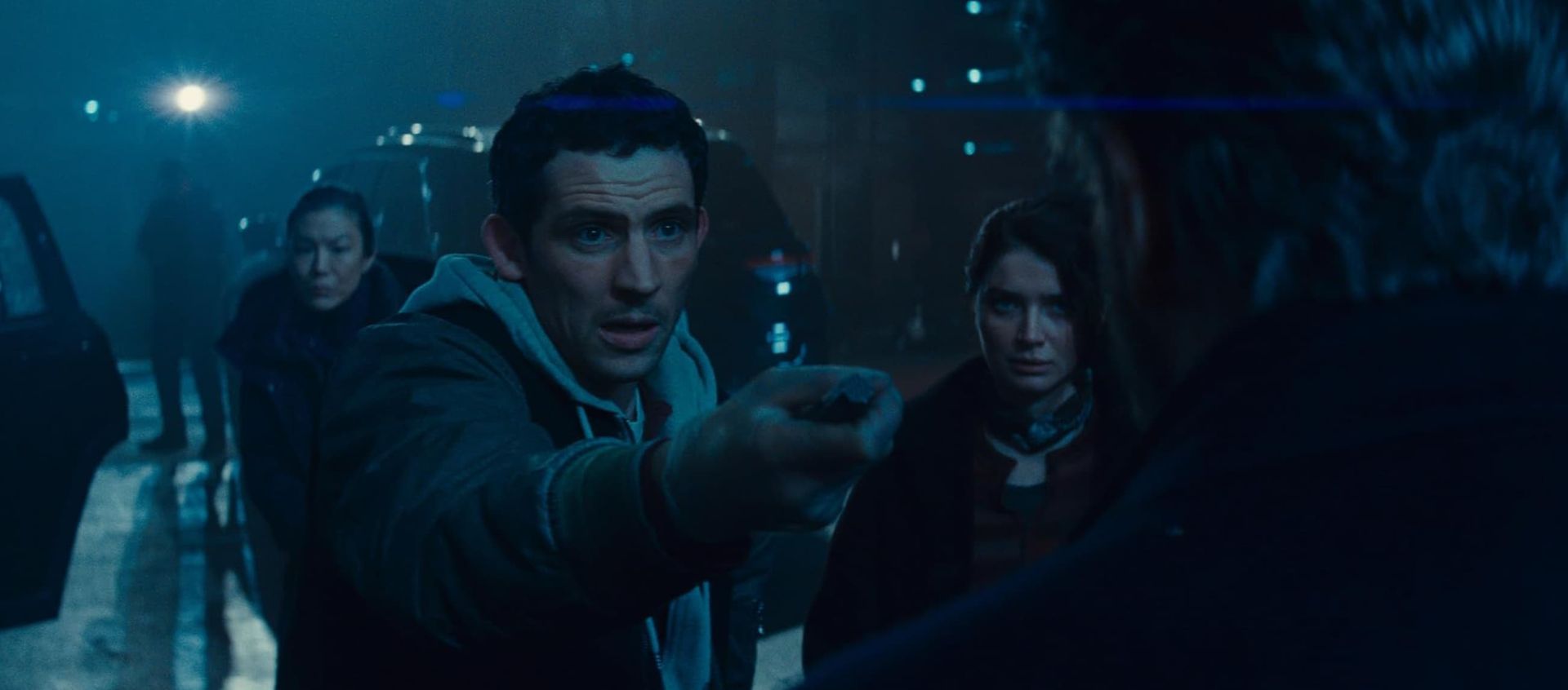 A tense nighttime scene with three people in a blue-lit forest, one pointing ahead.