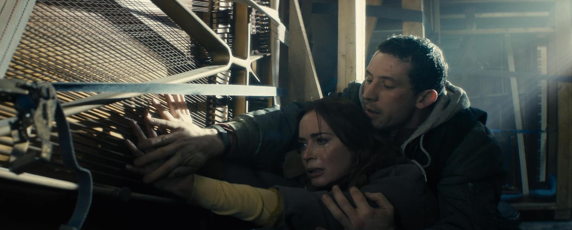 Two people crouch in a dim industrial space, shielding themselves near metal shelves as light cuts through dust.