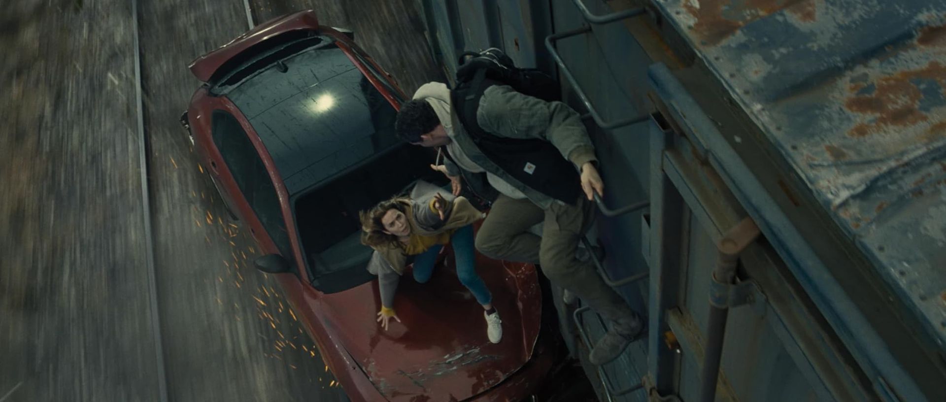 Two people crouch over an open red car in a narrow alley, one holding a flashlight.
