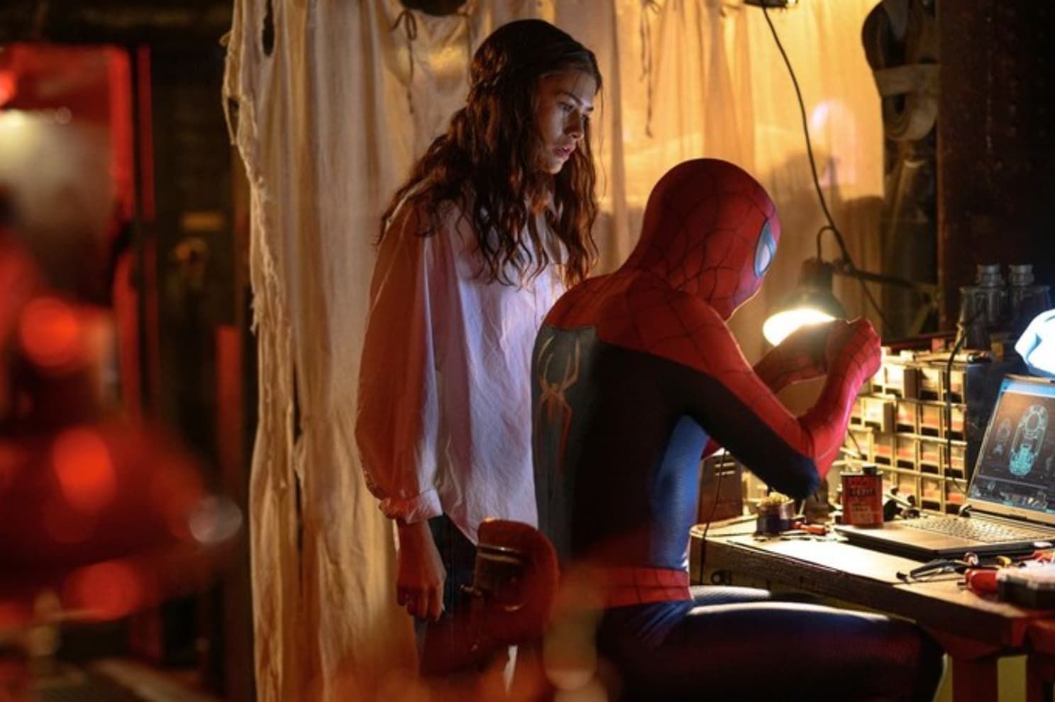 Masked person at a lit workbench while another stands behind, in a dim room with warm lights.