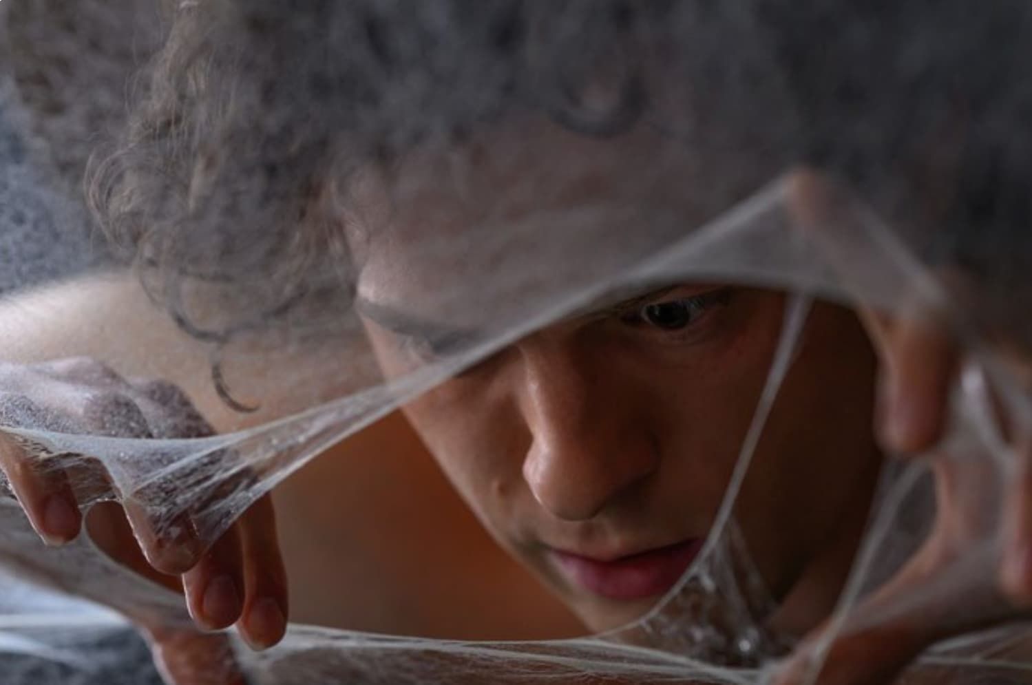 Peter Parker peering through a sheer fabric, hands holding it aside, with a focused expression