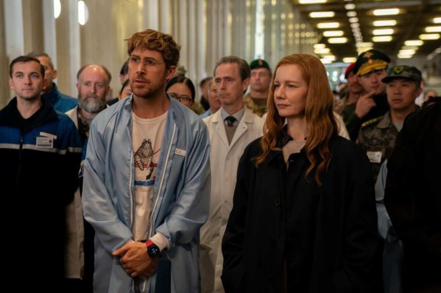 A group of people standing indoors, with a man in a light blue shirt and a woman in black in the foreground.