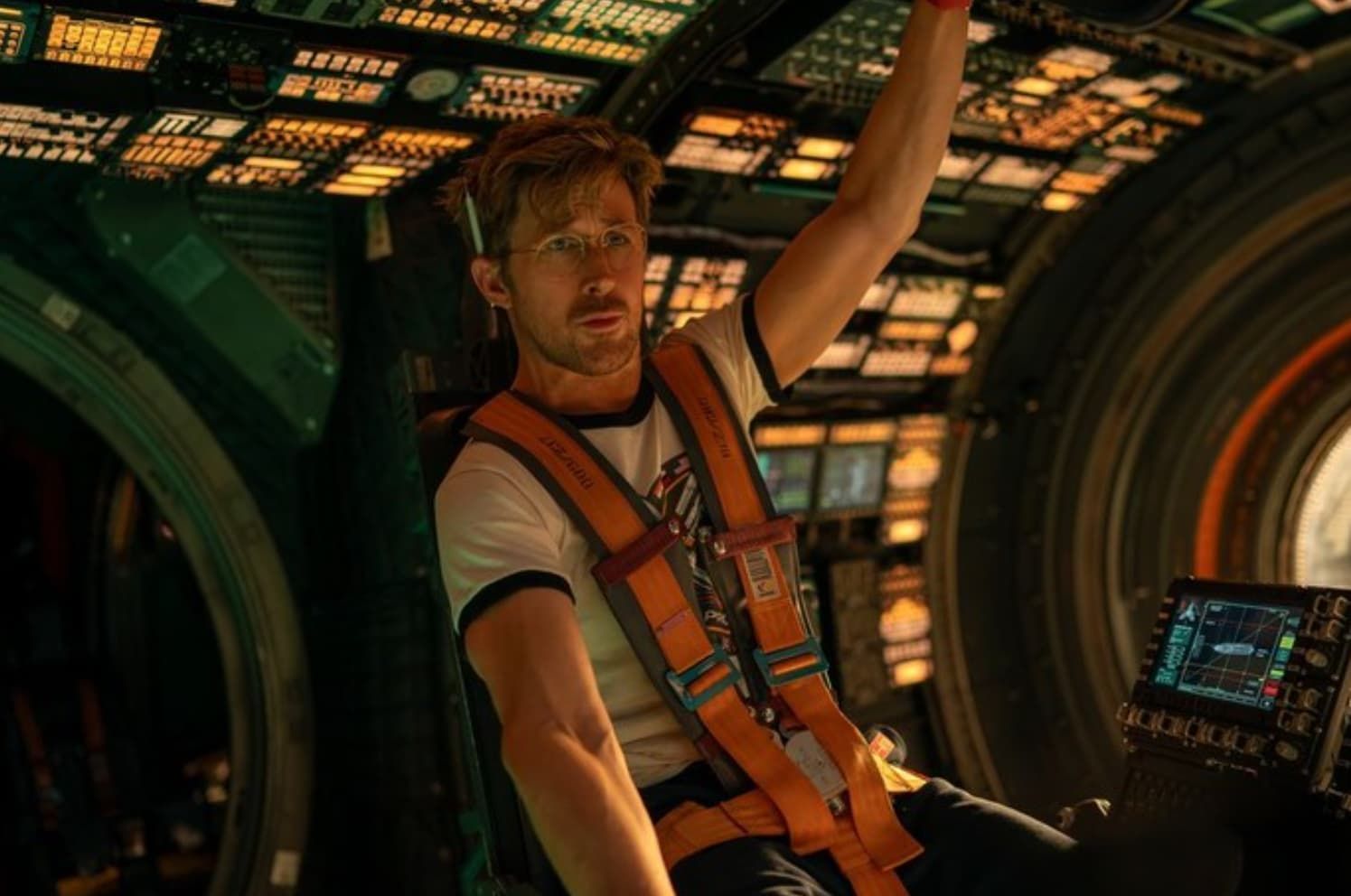 Man in an orange harness inside a spaceship, reaching up amid glowing control panels.