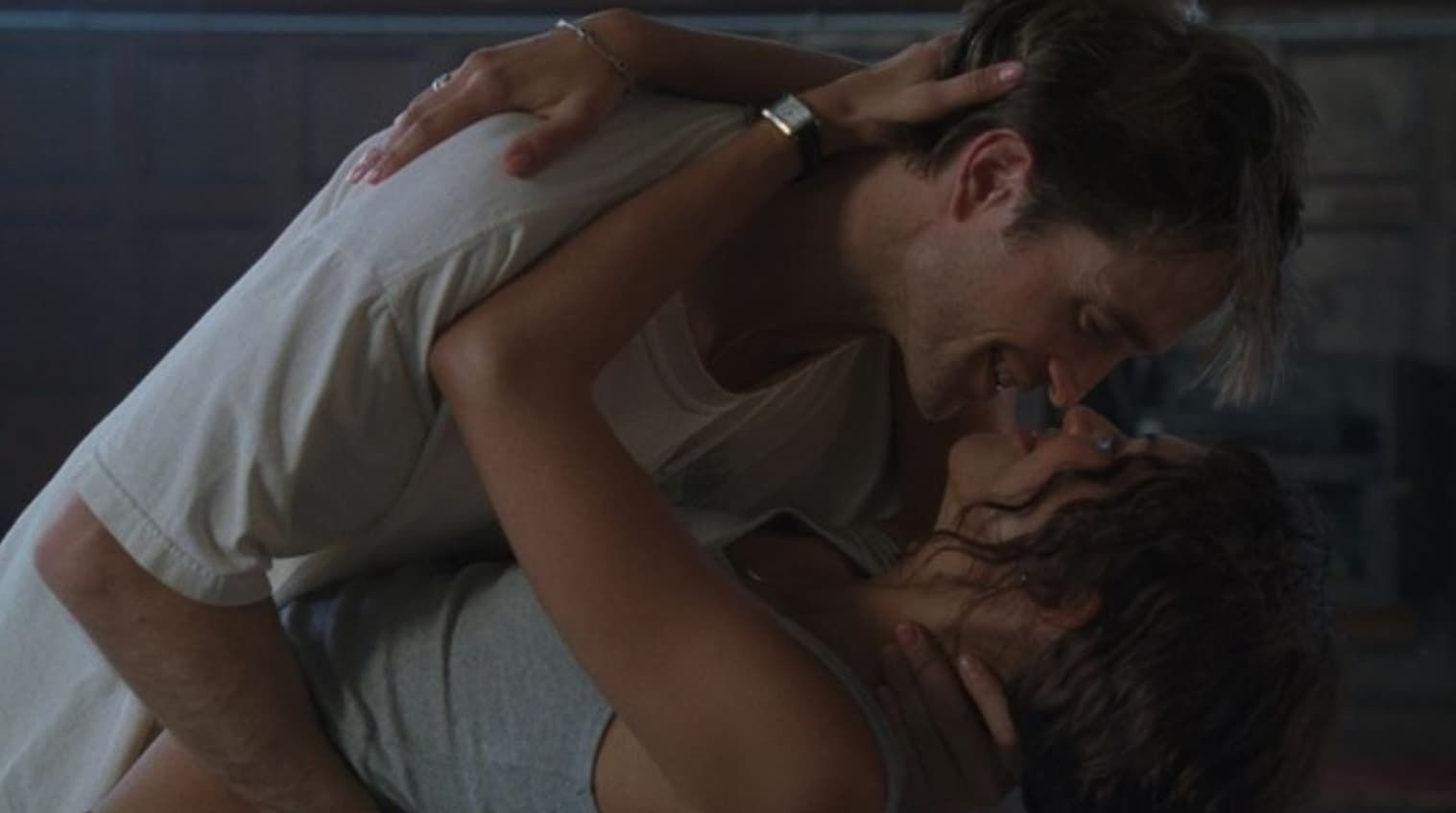 Two people kissing on a bed in a dimly lit room, one leaning over the other