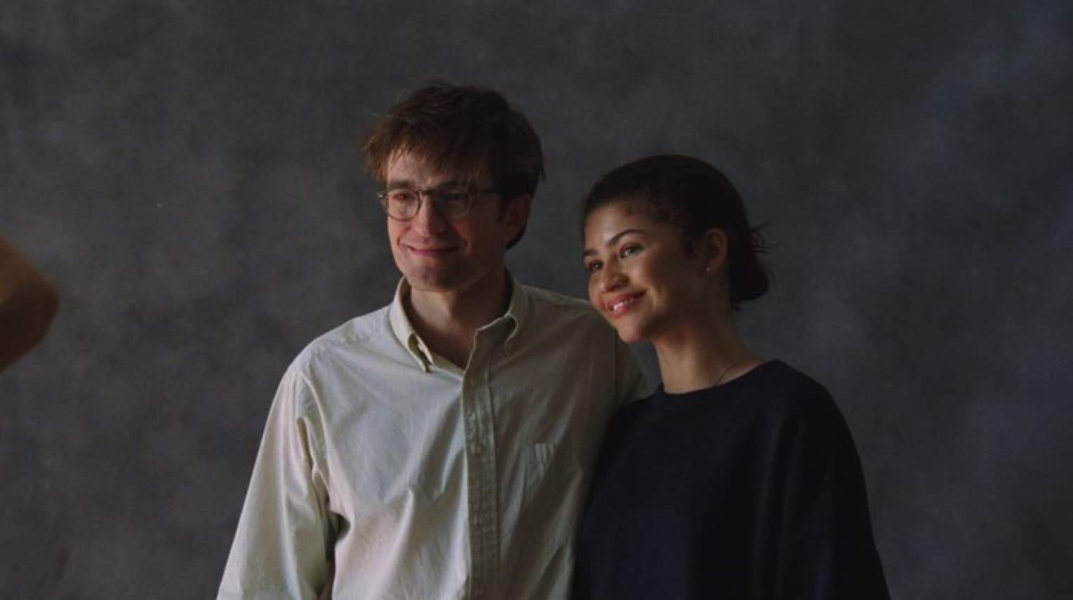 Two people smiling together in soft studio lighting, standing against a gray backdrop.