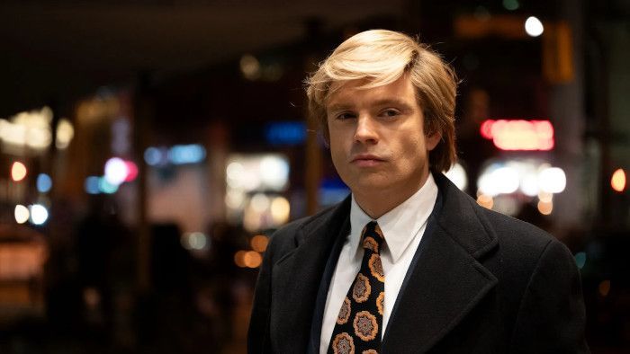Man with blonde hair in suit, looking forward, blurred city background.