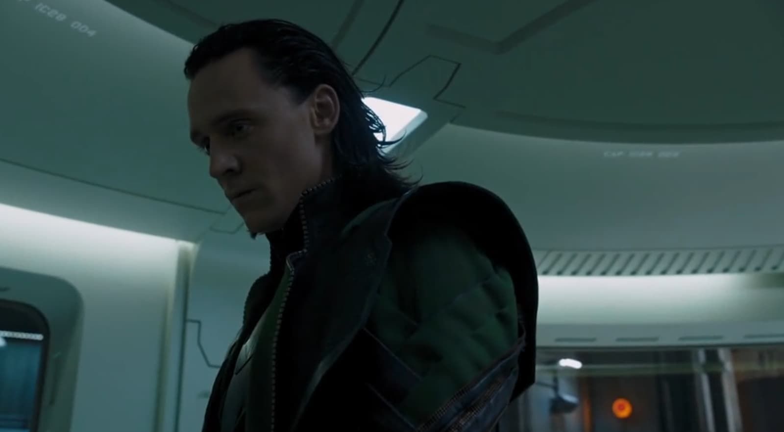 Dark-haired person in a dim sci-fi room, lit by greenish overhead lights, looking down pensively