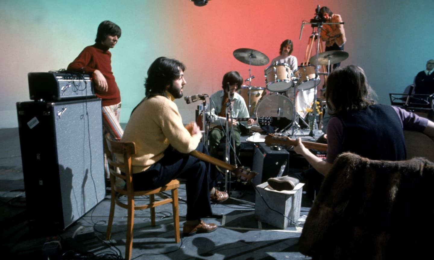 The Beatles rehearsing with guitars, drums, and cables under colorful pink-blue lighting.