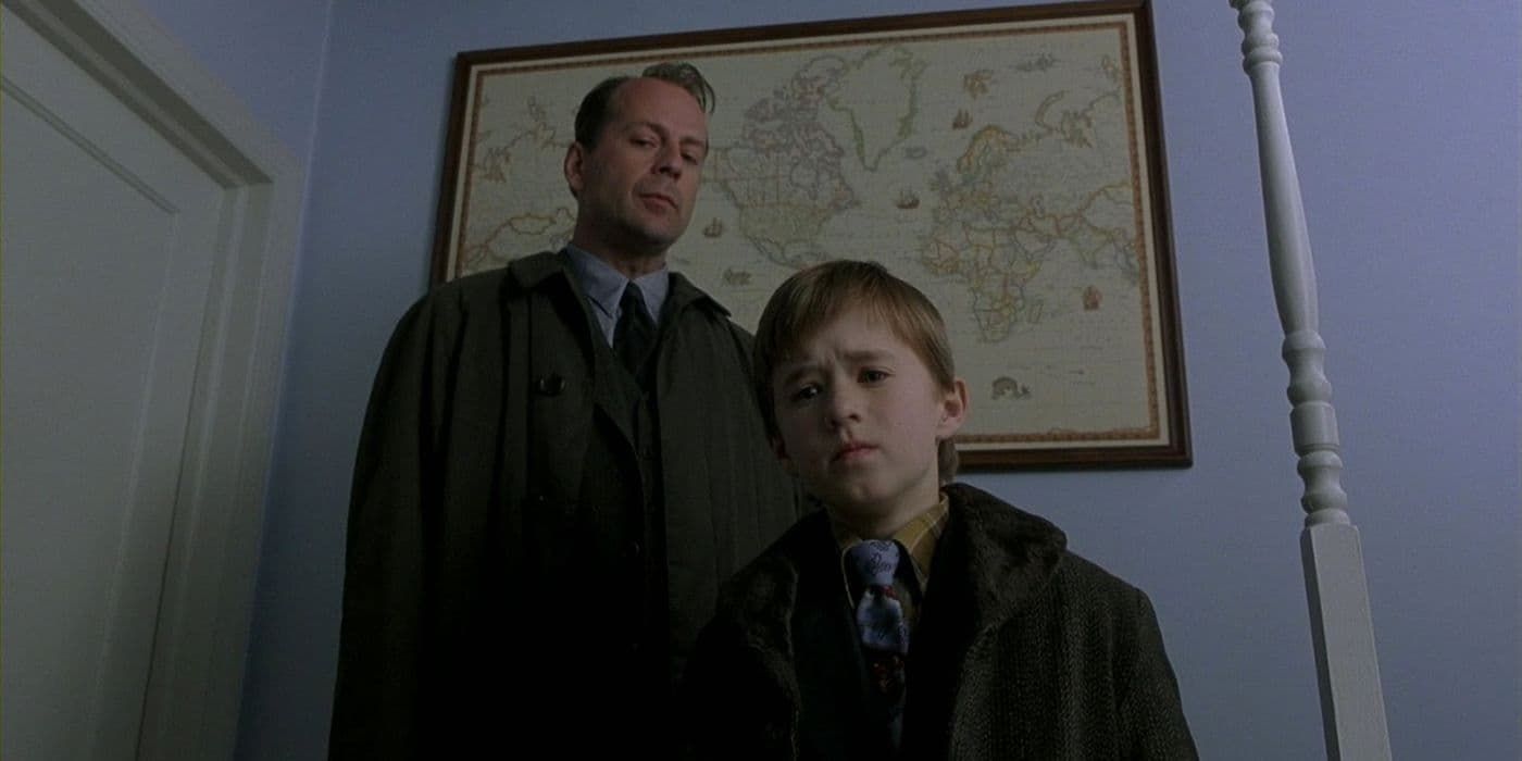 Two people stand indoors beneath a wall map, one in a dark coat and one in a suit.