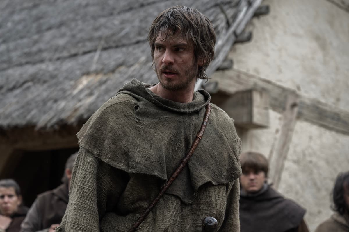 A young man in a rough green cloak stands in a muddy village, with thatched huts behind him.