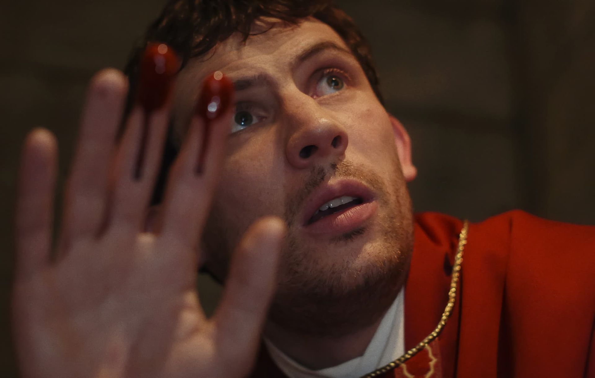 A man in a red robe raises a hand toward the camera, looking up with a worried expression.