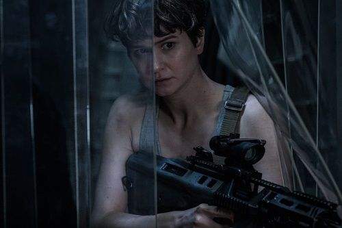 Woman in a tank top holds a rifle, looking out from behind a glass-paneled structure.