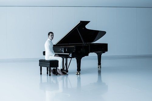 Pianist seated at a grand piano in a minimalist white room, playing. The piano is black.