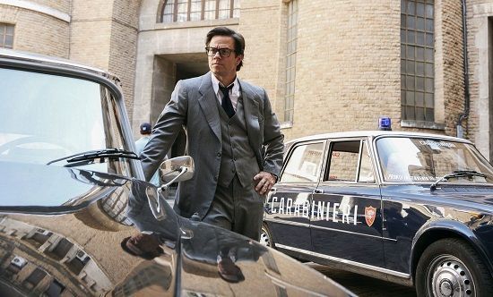 Man in suit leans on a car near a police car in front of a building.