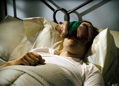 Man sleeping in bed with a sock covering his eyes. White shirt, gray comforter, dark headboard.