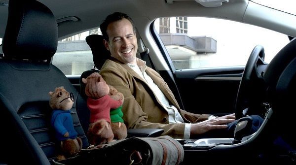 Man smiles in car, with Alvin and Simon chipmunk dolls in the front seat.