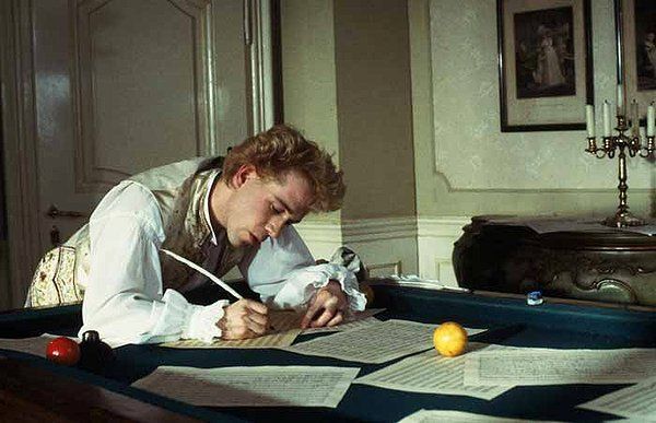 Man writing with a quill at a desk. He wears a white shirt and vest, in a dimly lit room.
