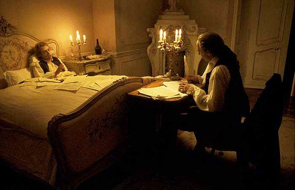 Man writing at desk beside a person in bed in a dimly lit room; candles illuminate the scene.