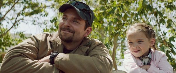 Man with beard in cap smiles next to a young girl. They sit outside near green foliage.