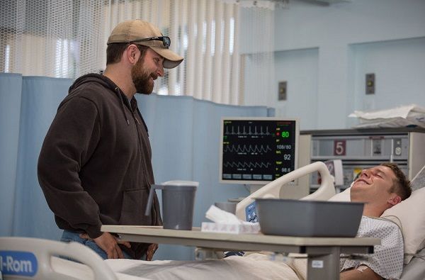 Man in brown hoodie smiles at patient in hospital bed; medical equipment visible.