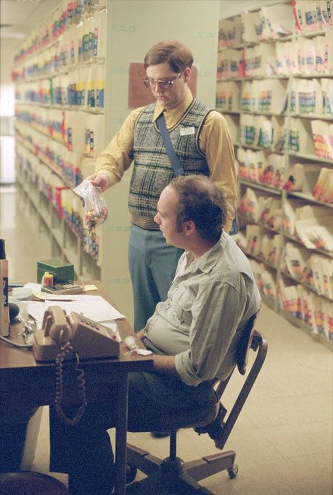 Two men in an office with files. One hands papers to the other at a desk, surrounded by shelving.