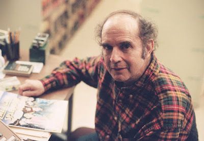 Man with thinning hair, plaid shirt, seated at a desk, looking at the camera.